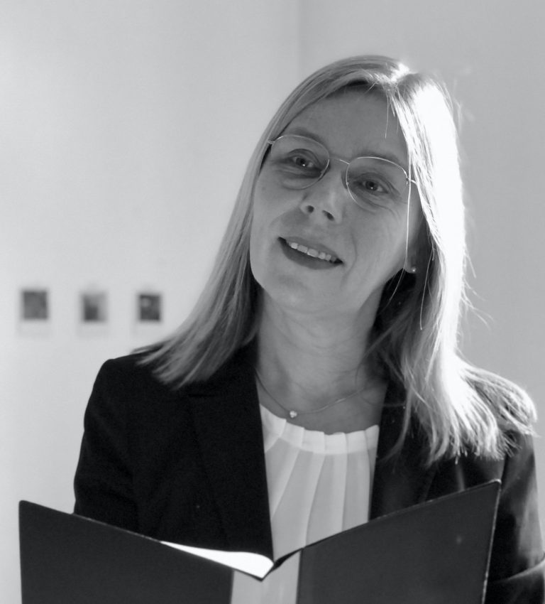 Frau mit blonden Haaren trägt einen Blazer und liest aus einer Textmappe vor.