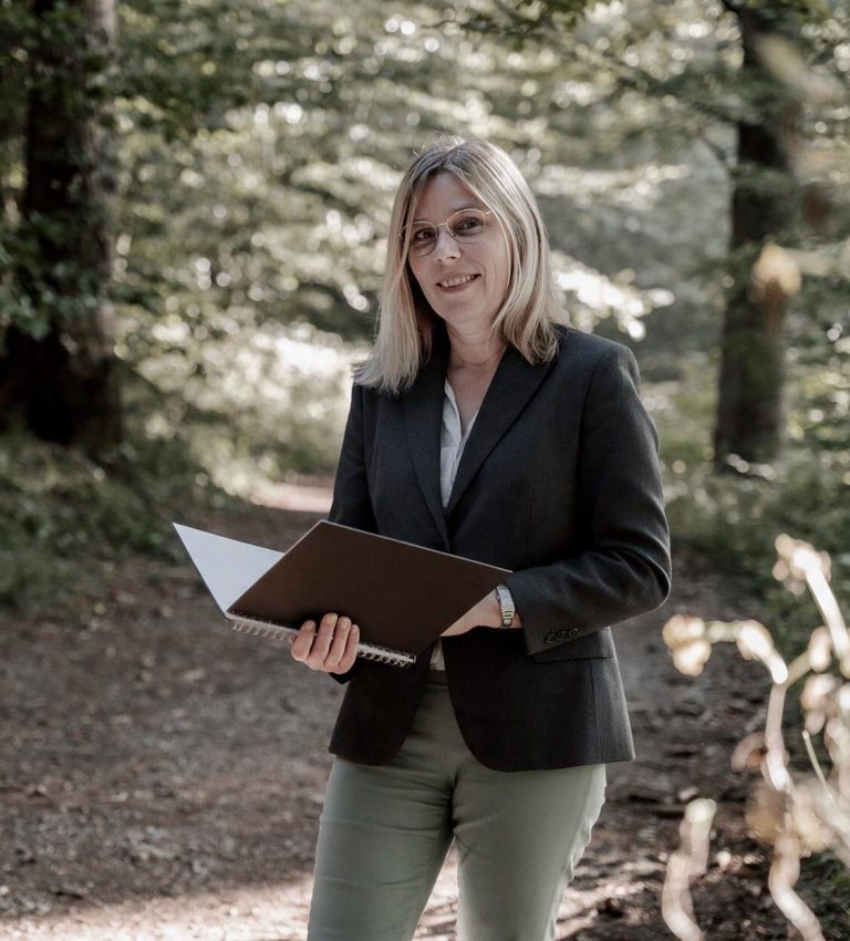 Frau mit blonden Haaren trägt einen Blazer und liest aus einer Textmappe vor.
