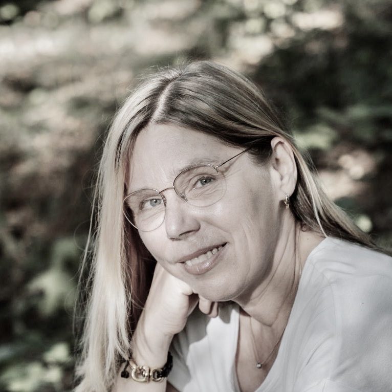 Eine Frau mit blonden Haaren und Brille lächelt sanft in die Kamera.