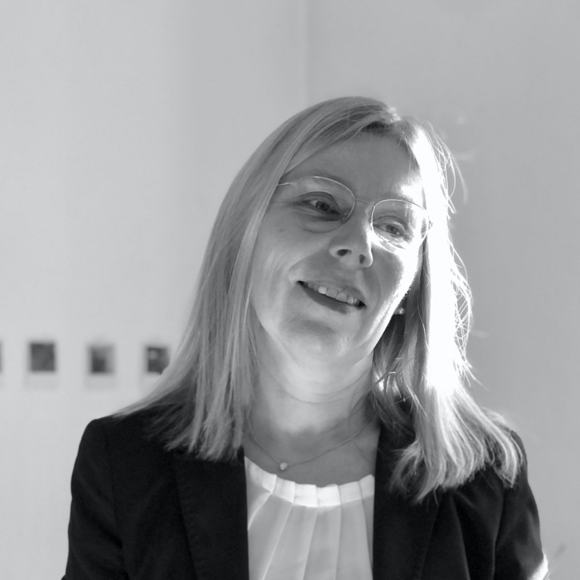 Frau mit blonden Haaren, in formeller Kleidung, lächelnd und nachdenklich.